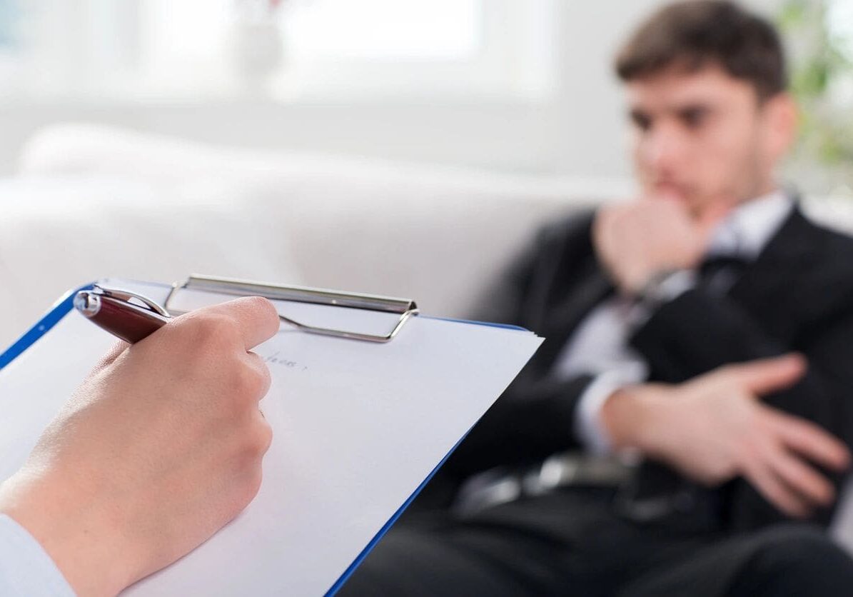 A person is writing on paper while another person sits in the background.